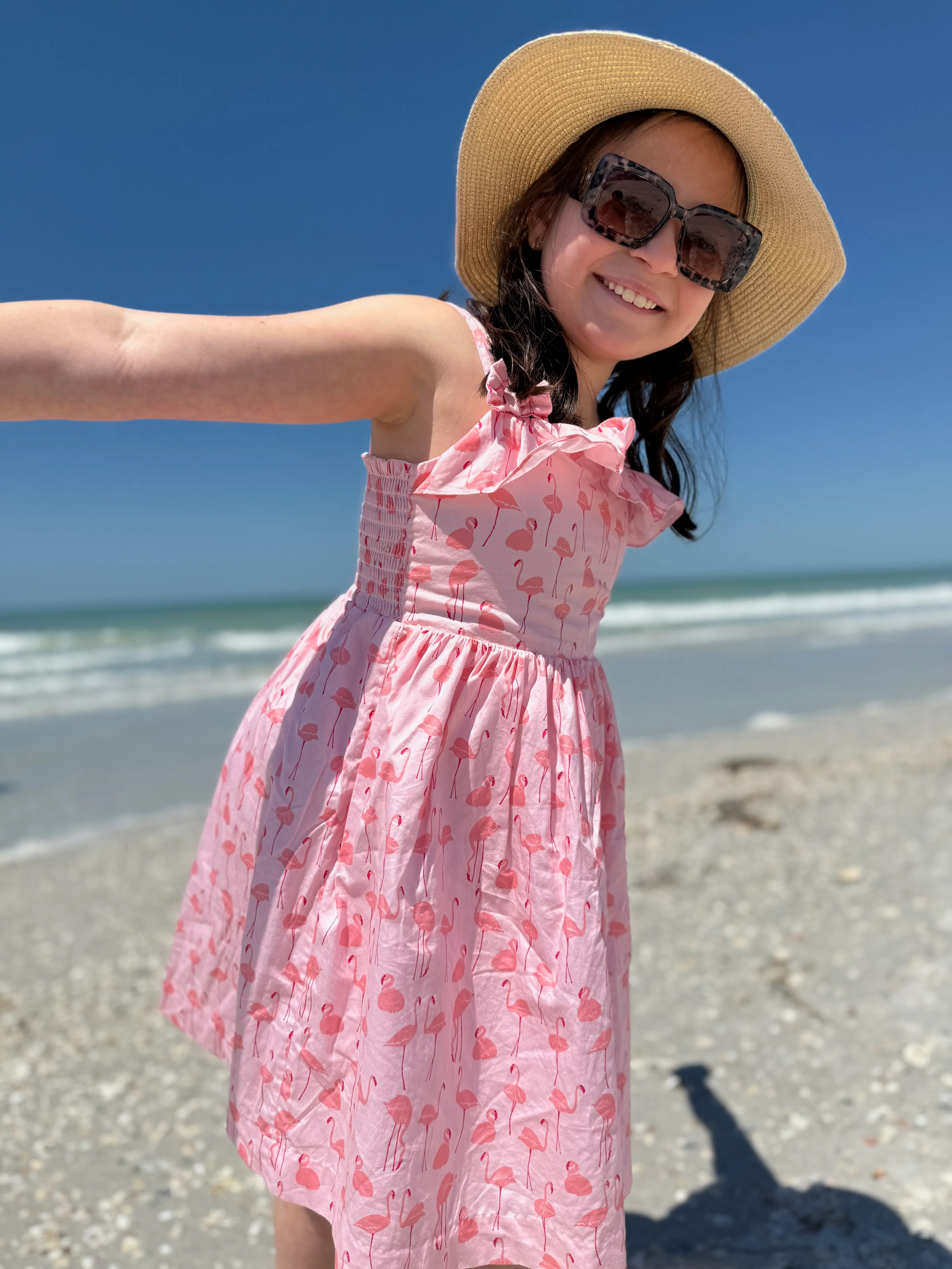 A smiling child in a flamingo-print sundress and sun hat on a bright Tampa Bay beach